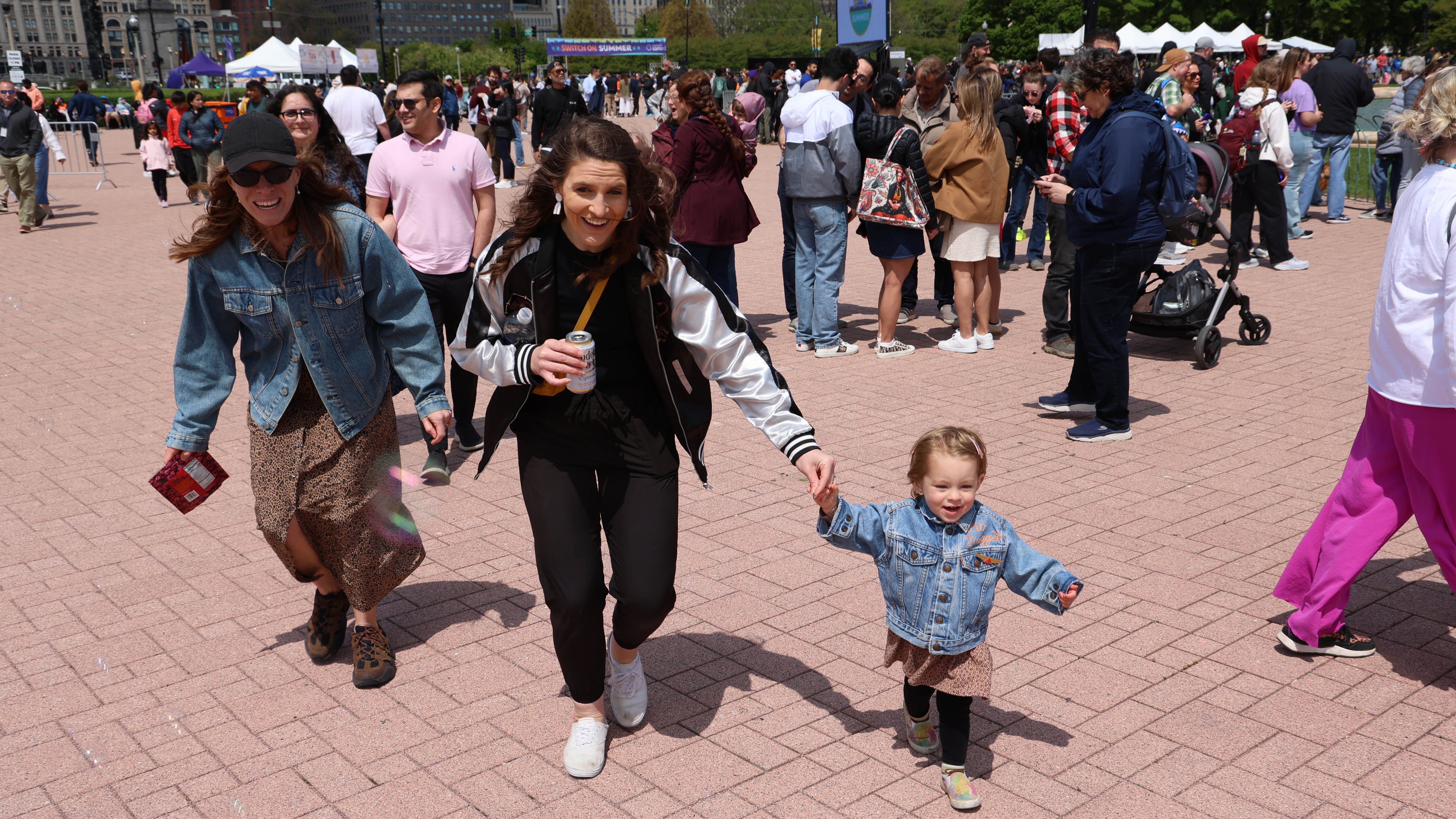 Two women and a child run across a paved area at a crowded outdoor event.
